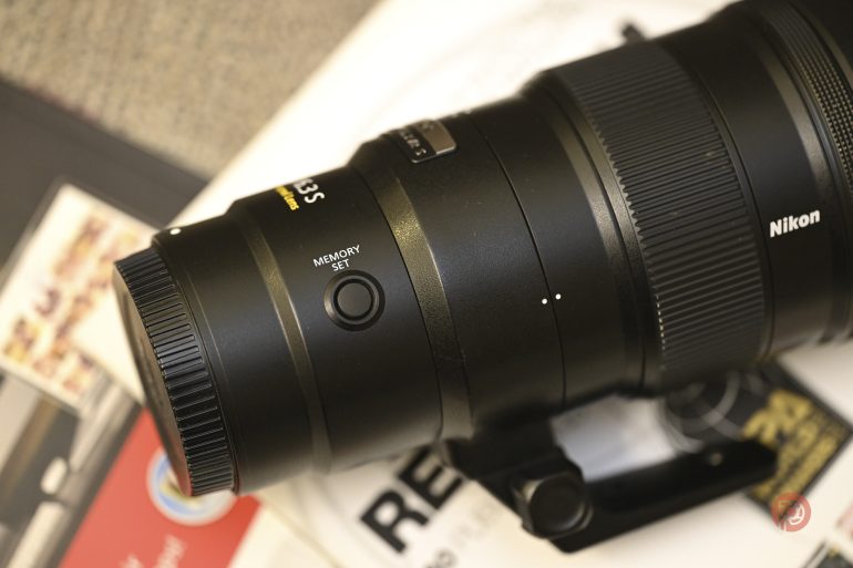 Close-up of a Nikon camera lens with a visible MEMORY SET button and part of its mount on a white surface.
