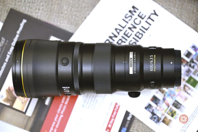 A Nikon Z 600mm f/6.3 S camera lens placed on top of magazines and documents on a light surface.