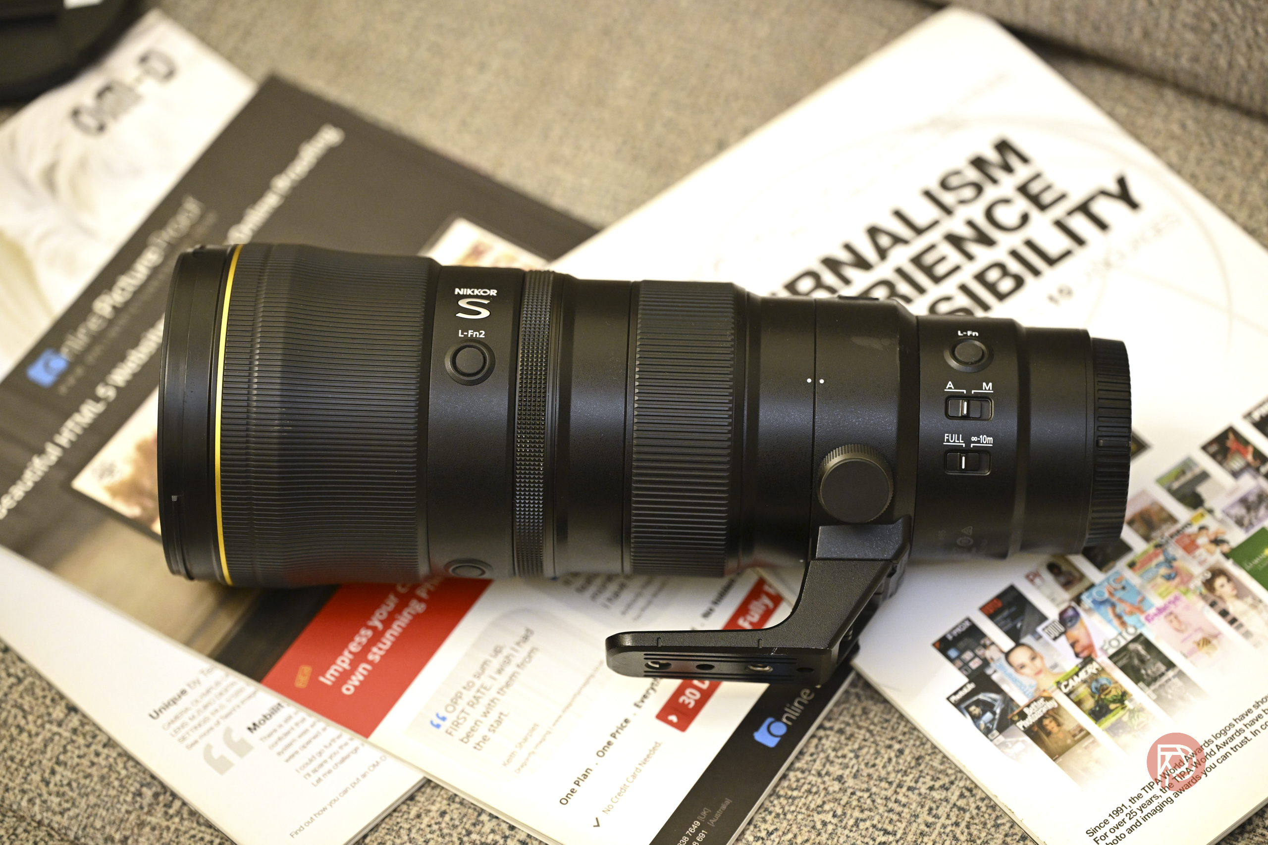 A Nikon telephoto camera lens rests on magazines and papers on a desk.