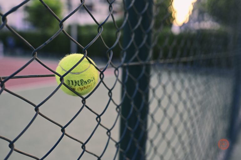 A tennis ball stuck in a chain-link fence on an outdoor tennis court.