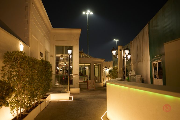 Outdoor restaurant patio at night, illuminated by warm lights and street lamps, with plants lining the walkway.