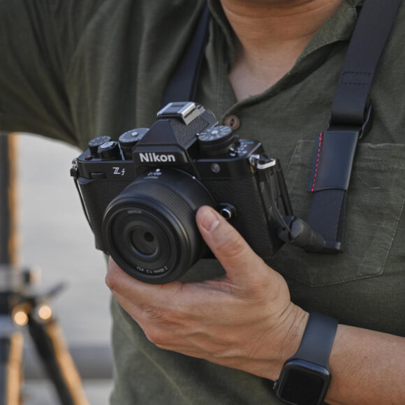 Person holding a Nikon Zfc camera with a wide-angle lens, tripod blurred in the background.