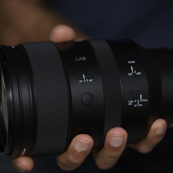 A hand holding a black camera lens with visible switches and textured focus ring.