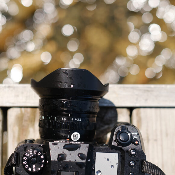 Close-up of a camera with a wet lens resting on a wooden surface, blurred water in the background.