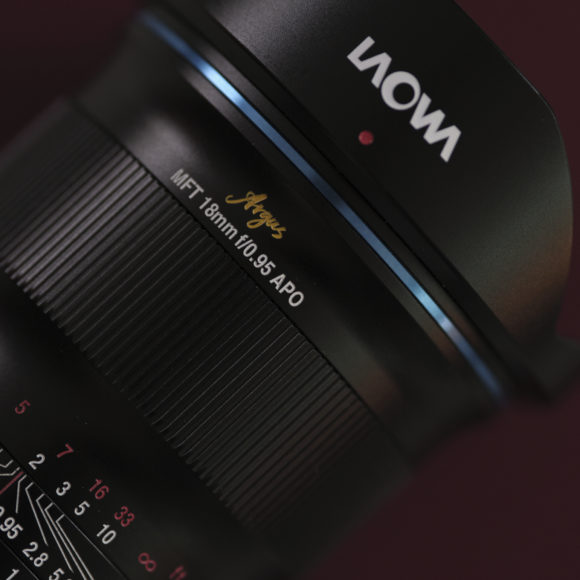 Close-up of a Laowa Argus MFT 18mm f/0.95 APO camera lens against a dark red background.