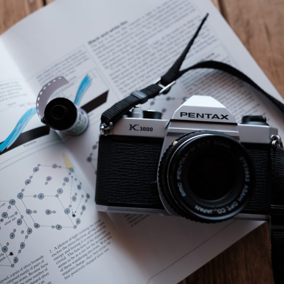 A Pentax K1000 camera and a film roll rest on an open book with diagrams, on a wooden surface.