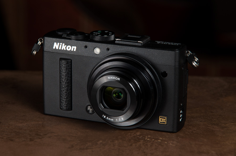 Black Nikon digital camera with a lens on a brown surface, angled slightly to the right.