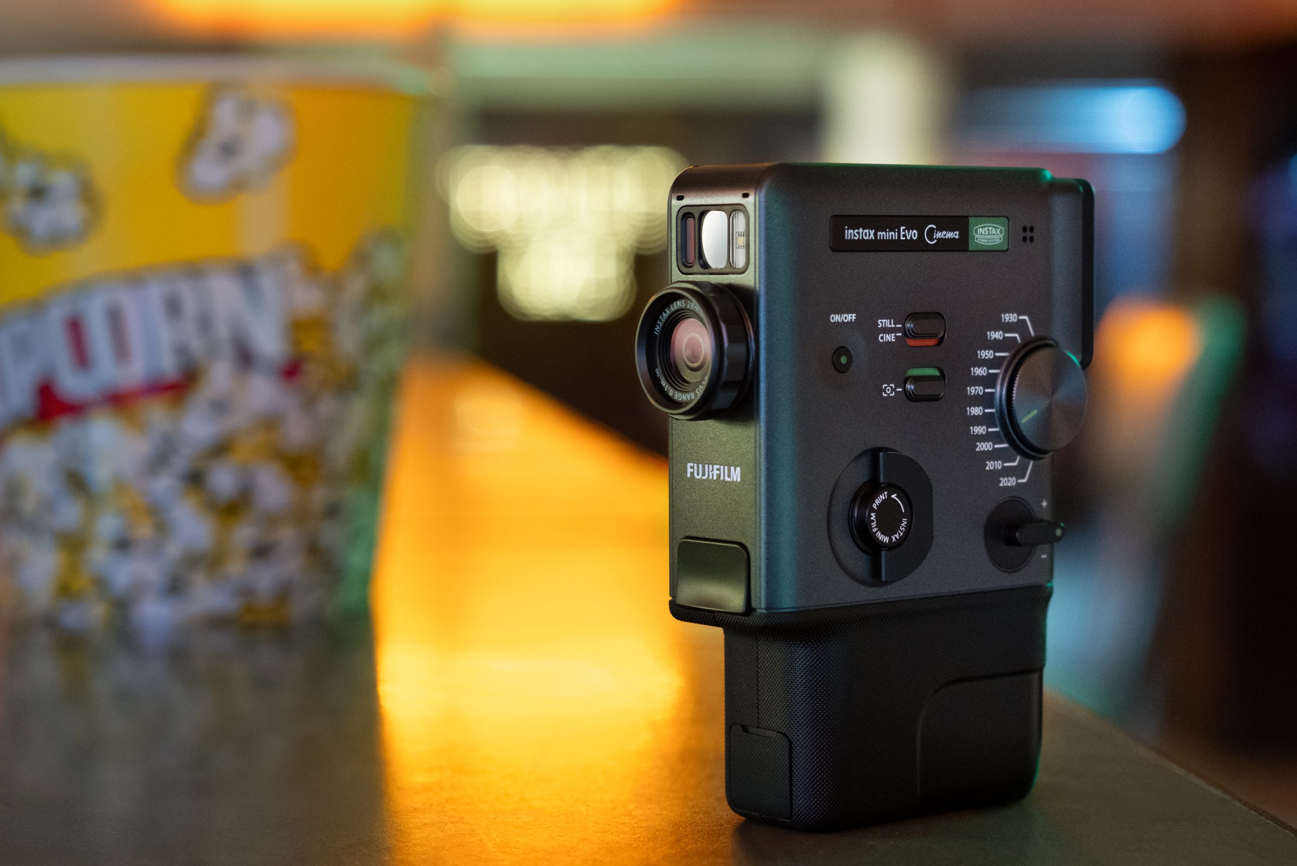 A Fujifilm Instax Mini Evo camera on a counter next to a yellow popcorn tub in a softly lit room.