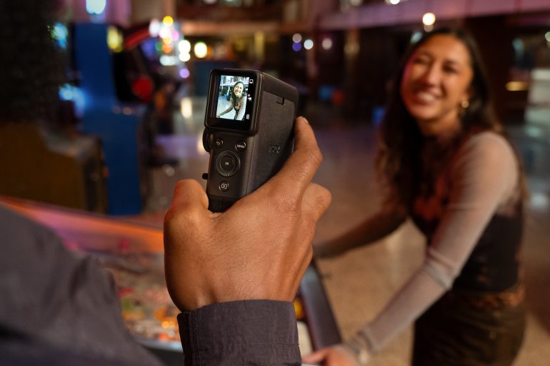 A person holds a camera filming a smiling woman near a pinball machine in a dimly lit arcade.