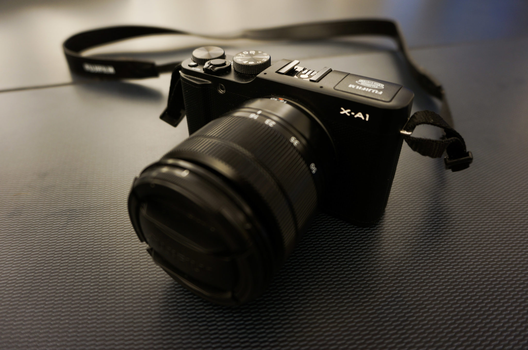A black Fujifilm X-A1 digital camera with a large lens rests on a textured dark surface.