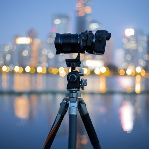 A camera on a tripod is set up by the water with a city skyline blurred in the background at dusk.