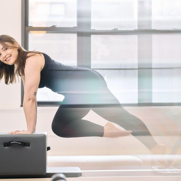 Woman in athletic wear smiling while balancing on a Pilates reformer in a bright studio.