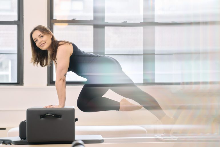 Woman in activewear doing a Pilates exercise on equipment in a bright studio with large windows.