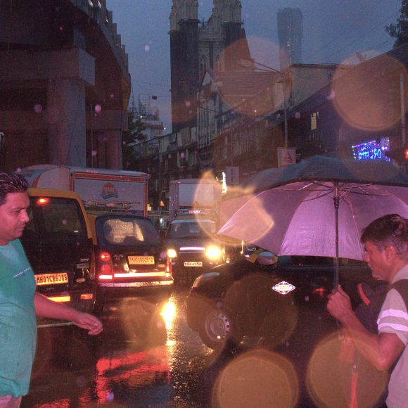 Two men walk on a busy, rainy street at night; one holds an umbrella, and traffic is visible behind them.