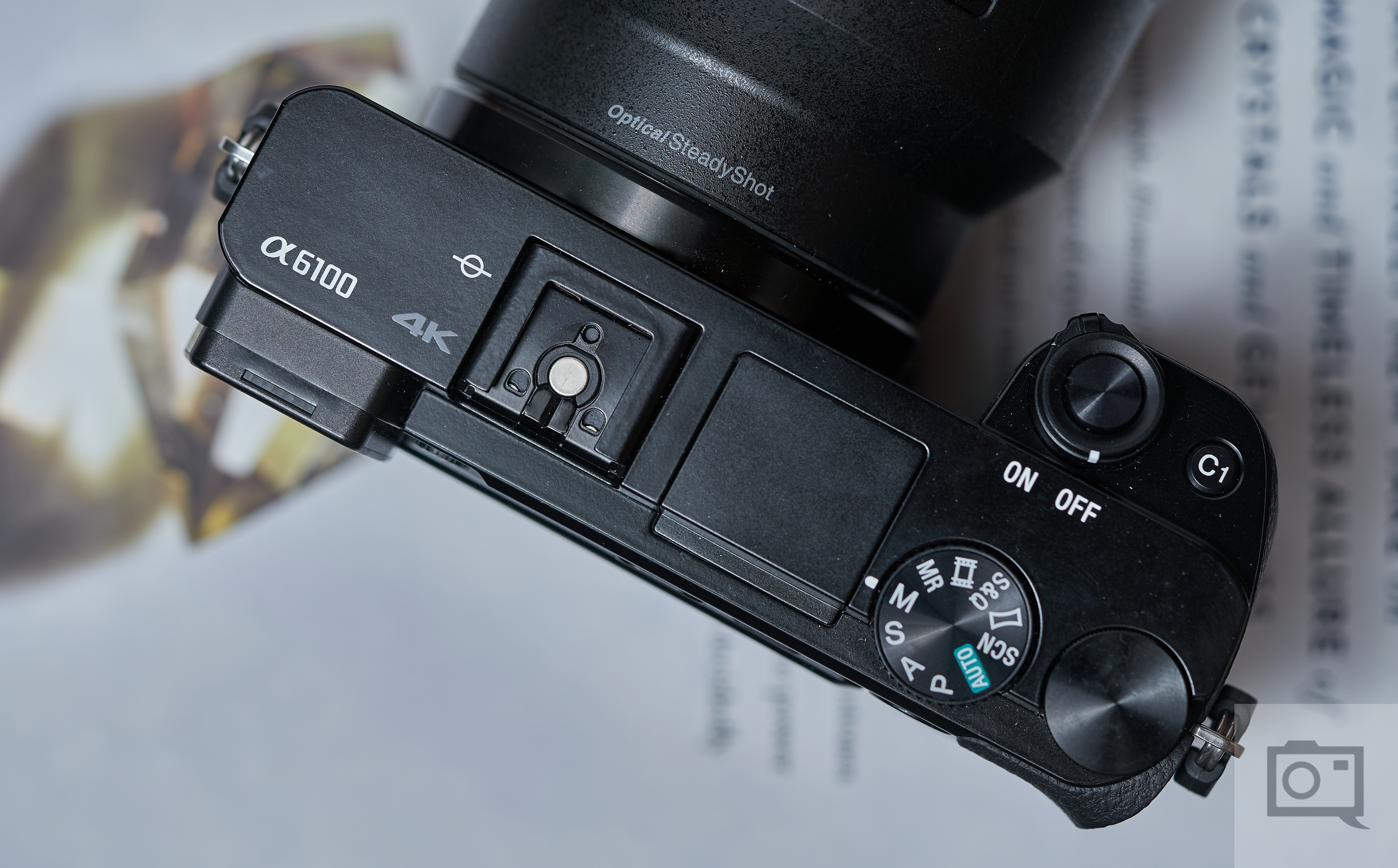 Top view of a Sony Alpha 6100 camera showing dials and controls, placed on a blurred magazine page.