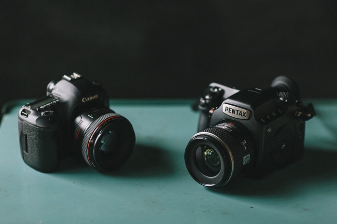 Two digital cameras, one Canon and one Pentax, placed on a teal surface with a dark background.