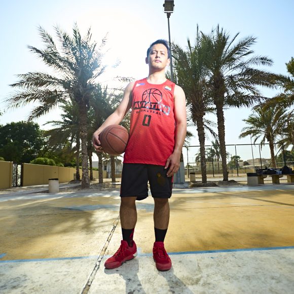 Person in a red jersey holding a basketball outdoors on a sunny court with palm trees.