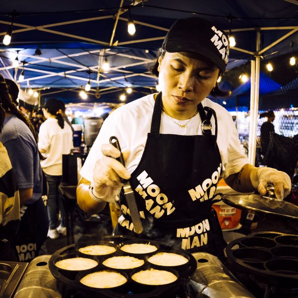 Person cooking pastries at a market stall under string lights, wearing a Moon Man apron and cap.