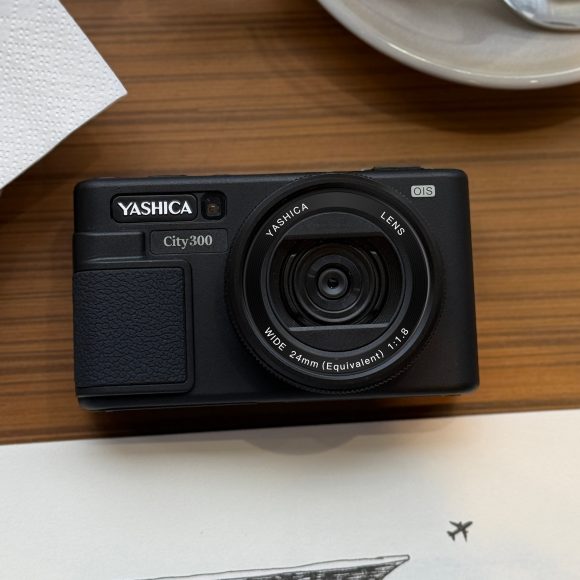 A Yashica City 300 camera on a wooden table near a napkin, plate, and a sketchbook.