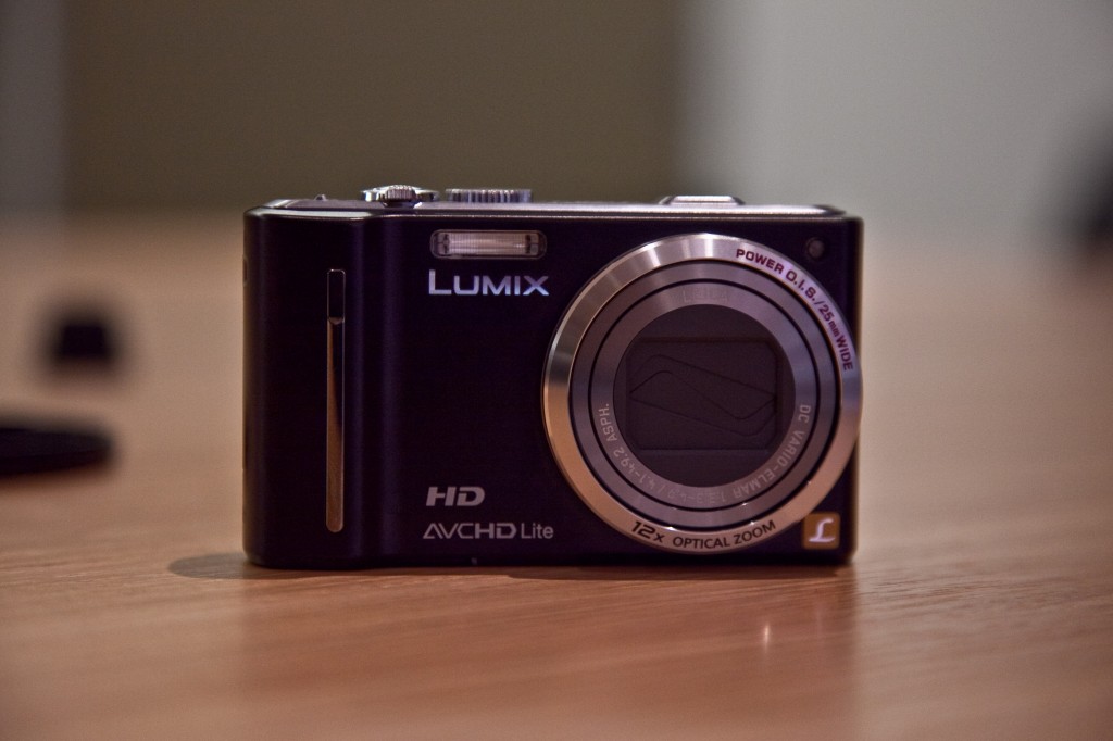 A black Lumix digital camera sits on a wooden surface with a blurred background.