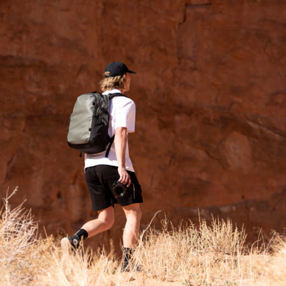 Person hiking in a desert landscape, wearing a WANDRD STRATUS 18L and holding a camera, with a rocky backdrop.