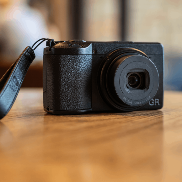 A compact black digital camera with a wrist strap rests on a wooden table indoors.
